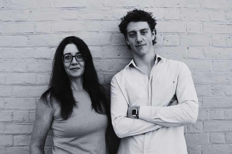 Two people standing against a white brick wall in black and white photo. Woman on left with long dark hair and glasses, man on right in white shirt with arms crossed.