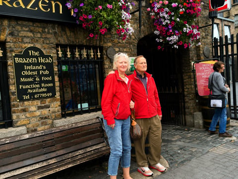 Two people in red jackets stand outside the Brazen Head pub in Ireland, with hanging flower baskets and stone architecture visible.