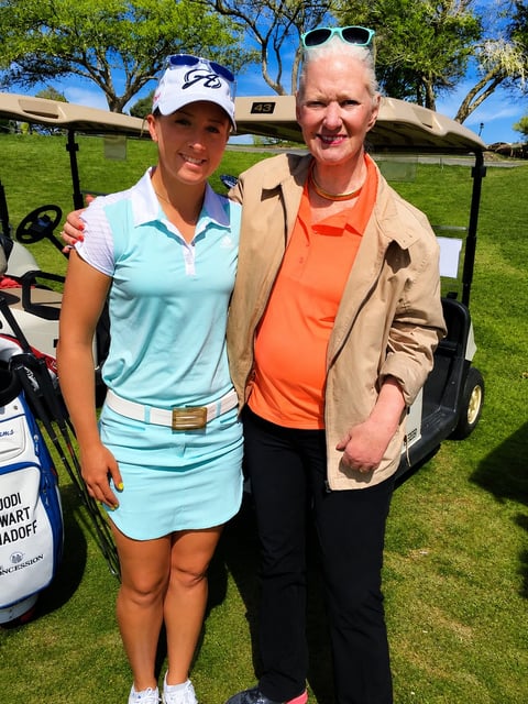 Two people posing together on a golf course. A woman in light blue polo outfit and white cap stands next to an older man in orange shirt and tan jacket with sunglasses.