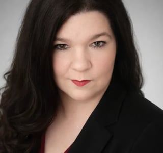 Professional headshot of a woman with long dark hair wearing a black blazer against a gray background