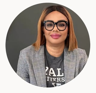 Portrait of Joy Nwabueze — a woman with reddish-brown hair, black-framed glasses, and pink lipstick wearing a plaid blazer over a dark graphic t-shirt, circular profile photo on a gray background