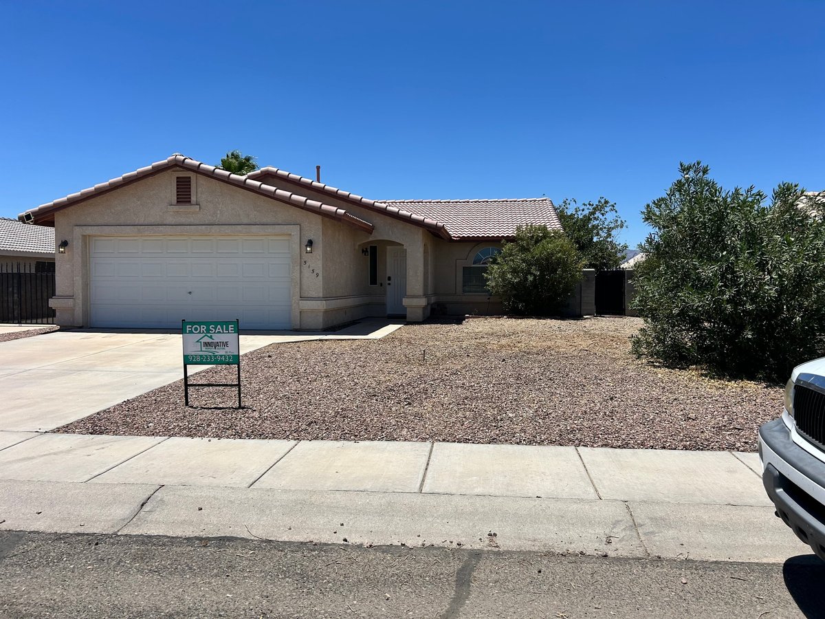Single-story beige house with white garage door, gravel front yard, and For Sale sign under clear blue sky
