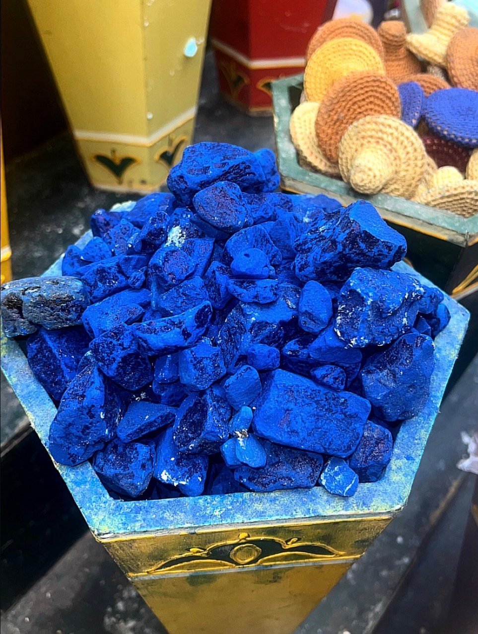 Bright blue azurite mineral chunks displayed on a decorative gold base, with other mineral specimens visible in the background
