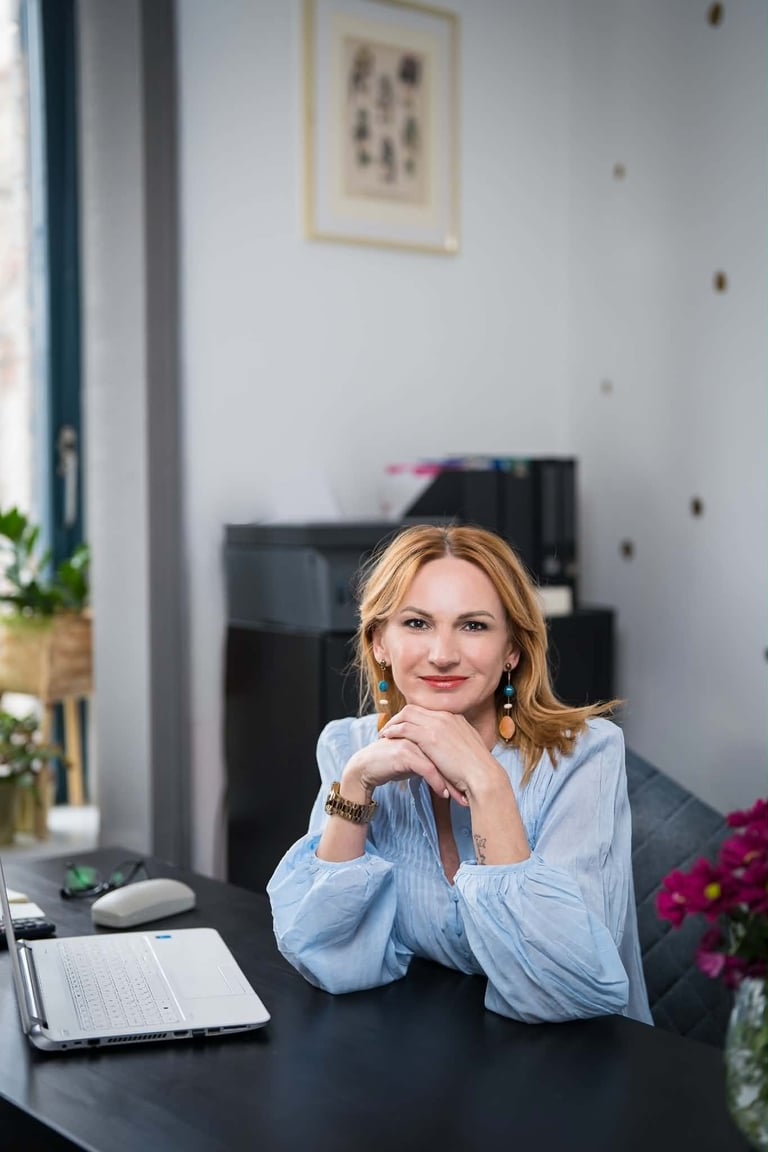 Woman with reddish-blonde hair in light blue shirt sitting at desk with laptop and flowers, smiling at camera in modern office