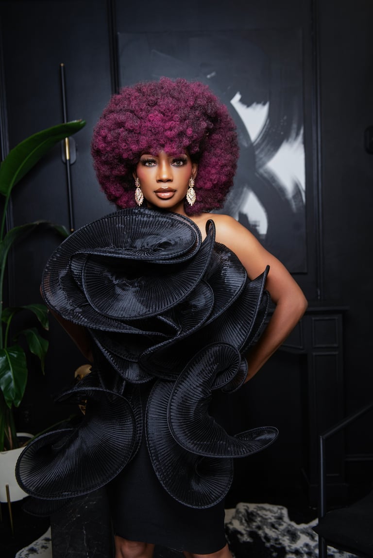 Woman with voluminous magenta afro wearing an avant-garde black sculptural outfit poses against a dark studio backdrop