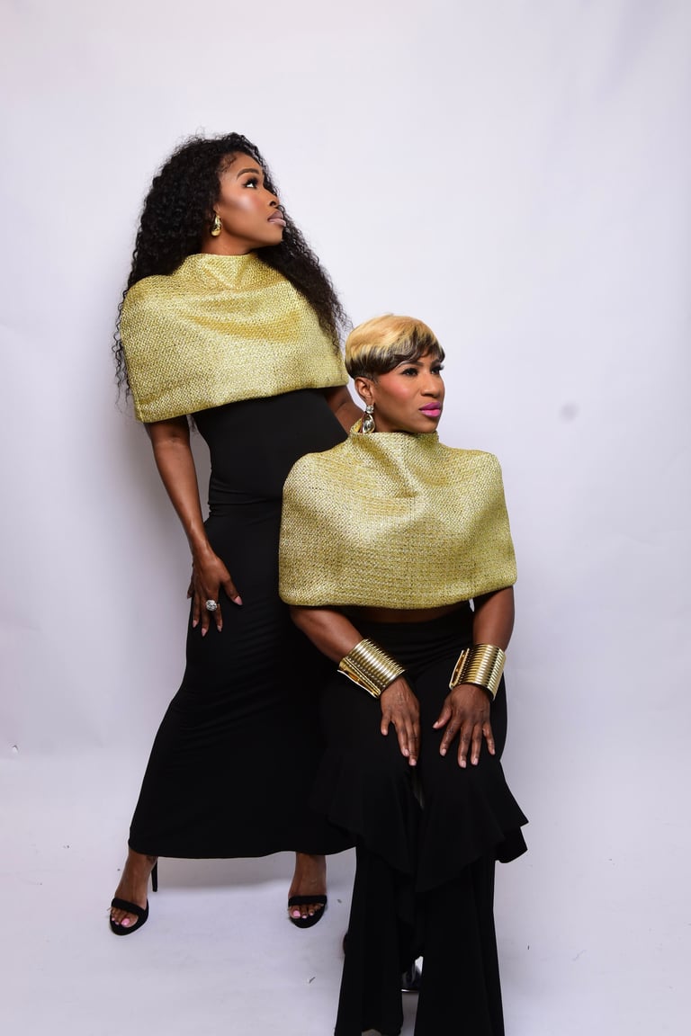Two women posing together wearing matching gold and black outfits with gold capes and accessories against a white background