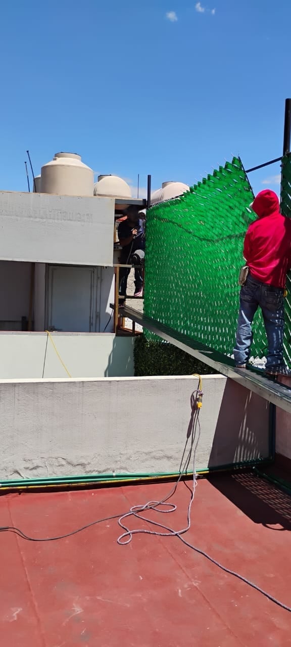 Construction workers in red jackets on a rooftop with industrial equipment and green safety netting under clear blue sky