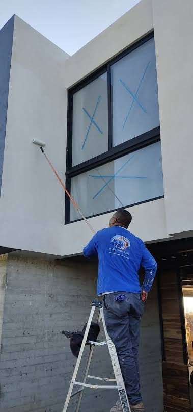 Worker in blue shirt on ladder cleaning large window with X marks on modern white building
