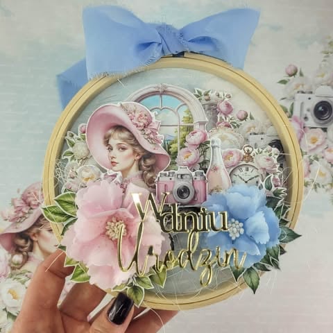 Hand holding circular vintage-style scrapbook embellishment featuring a woman in pink hat with flowers, cameras, and blue ribbon bow