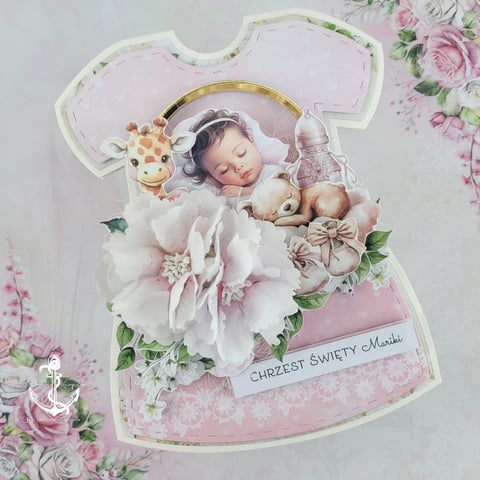 Decorative baby girl card shaped like a dress with sleeping infant, flowers, toys, and text reading Święty Chrzest
