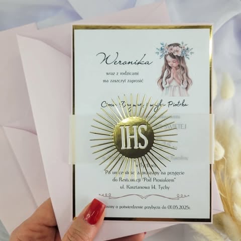 Hand holding a white invitation card with decorative gold border, gold sunburst IHS symbol, and 'Weronika Cioci Bogumiły i wujka Piotra' Polish text for First Communion