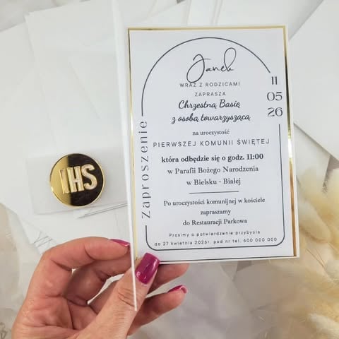 Hand holding gold IHS religious pin badge next to white wedding invitation card 'Janek Chrześcia Basie z osobą towarzyszącą' with decorative gold border