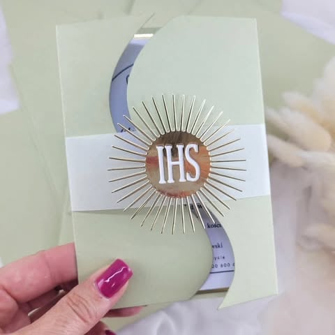 Hand holding a cream-colored invitation card with gold sunburst IHS design and sage green ribbon band