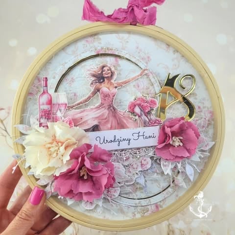 Decorative embroidery hoop with illustration of woman in pink dress, surrounded by cream and magenta artificial flowers, gold 18 numerals, and 'Urodziny Hani' text