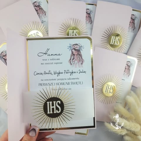 Religious invitation cards with gold IHS symbol and decorative sun rays, displayed in hand. 'Hanna Cioci Anety, Wujka Patrycja i Julii' Polish text for First Holy Communion