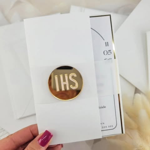 Hand holding a white invitation card with gold IHS religious pin badge 'Dominika' against blurred background of documents