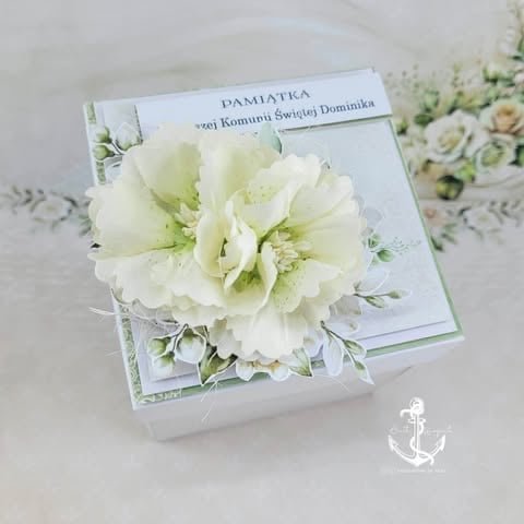 White decorative box with cream and white flowers and greenery, topped with a floral greeting card on a light background.