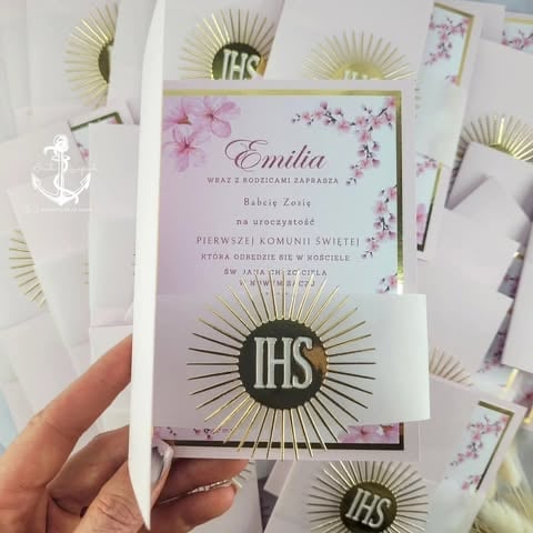 Hand holding a religious invitation card with pink cherry blossom floral design, gold radiating IHS symbol, and 'Emilia Babci Zosi' Polish text for First Holy Communion