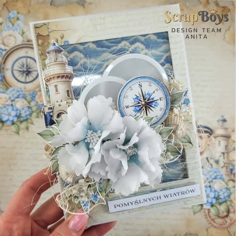 Hand holding a handmade scrapbook card featuring a white flower, compass rose, lighthouse, and nautical design elements.