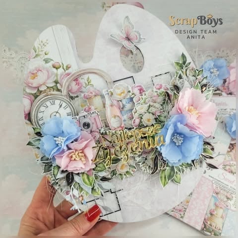 Hand holding a decorative white scrapbook card with pink and blue 3D flowers, clock, butterfly, and golden 'W dniu Urodzin' text embellishments