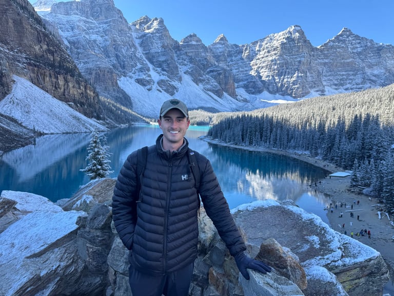 Andrew Donohue on the rocky shore of an alpine lake with mountains and forest in background