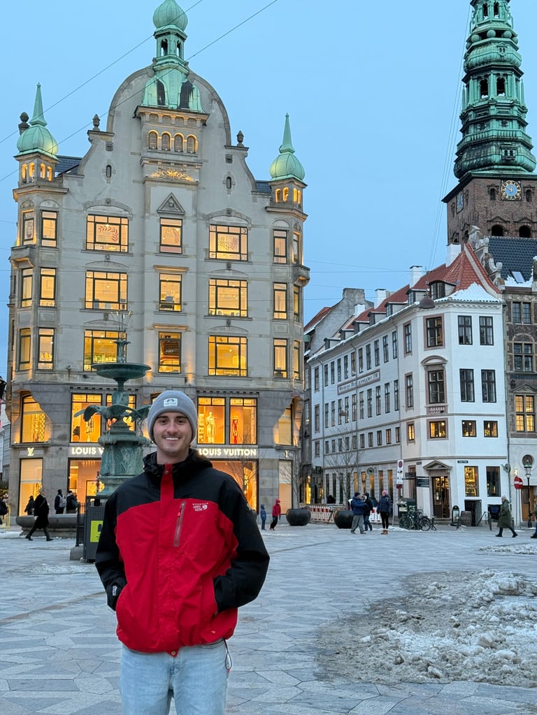 Andrew Donohue in red jacket and beanie smiling in a European city square with historic buildings