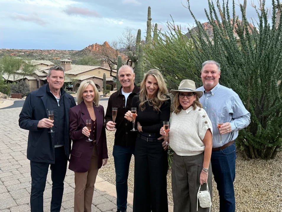 Six people standing together outdoors in a desert landscape, holding drinks and smiling at the camera