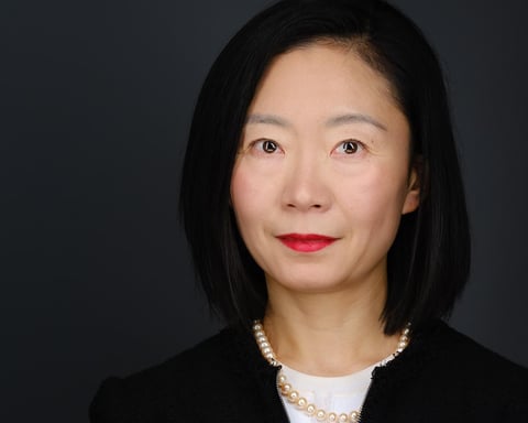 Professional headshot of an Asian woman with shoulder-length black hair, wearing red lipstick, a black blazer, and pearl necklace against a dark background.