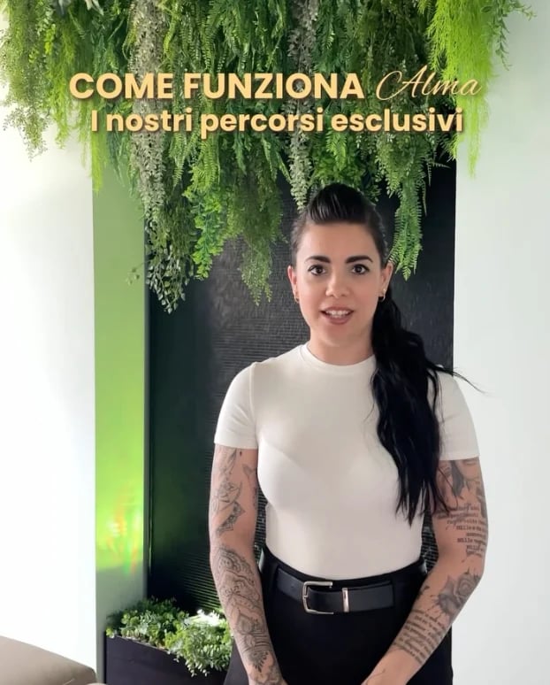 Woman with long dark hair and arm tattoos wearing white t-shirt and black pants, standing under hanging green plants with yellow neon lighting