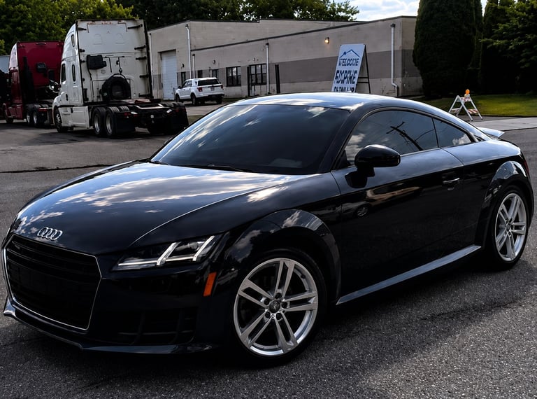 Black Audi sports car with silver alloy wheels parked in a lot with delivery trucks in background