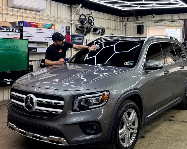 Technician applying protective film to the hood of a gray Mercedes-Benz SUV in a garage workshop
