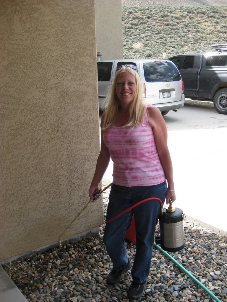 Comfort Pest Control owner Carol Westin, a woman with blonde hair in a pink tank top and jeans, holding a professional pest control sprayer and spray wand, standing beside a residential concrete column