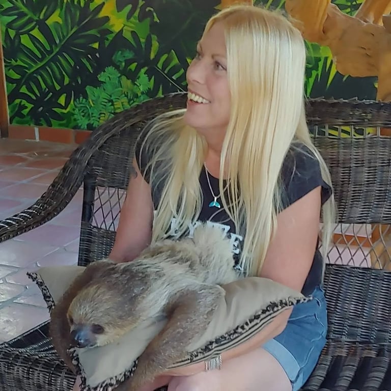 Woman with blonde hair smiling while holding a sloth on her lap, with tropical plants in the background