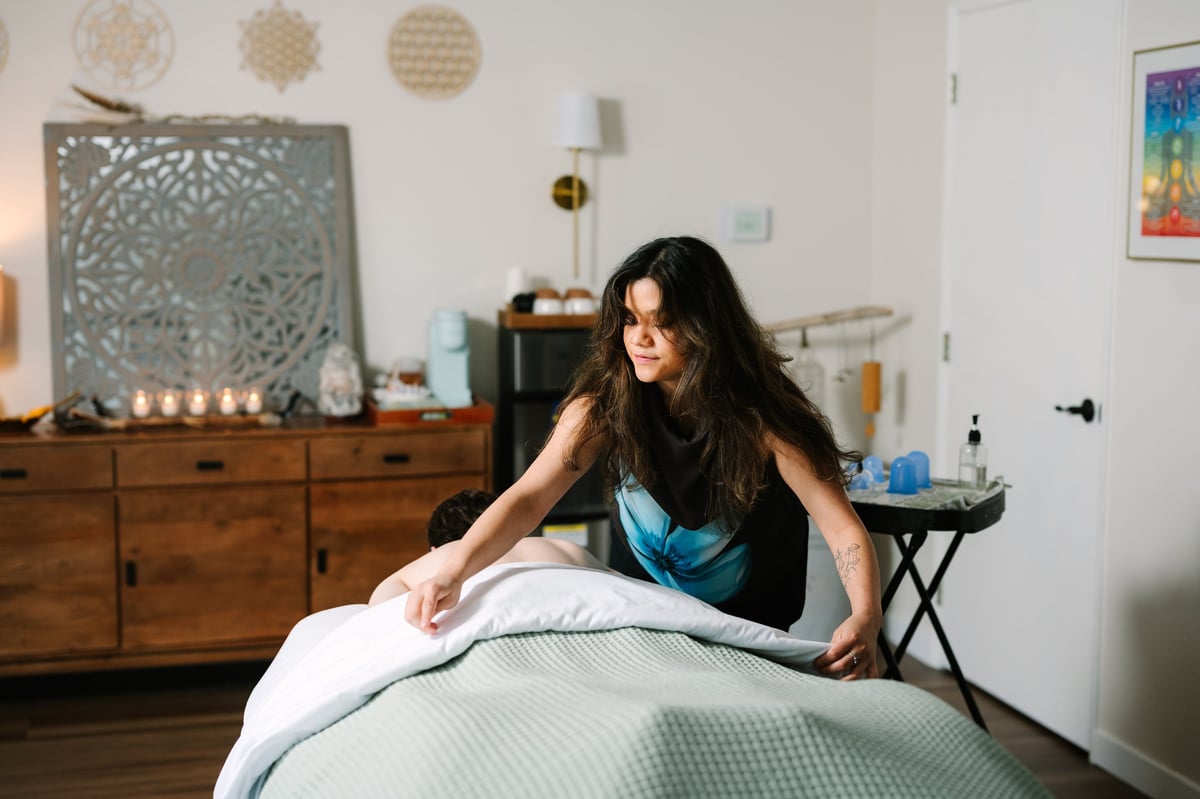 Woman with long dark hair sitting on a bed in a bedroom with wooden furniture and decorative wall art