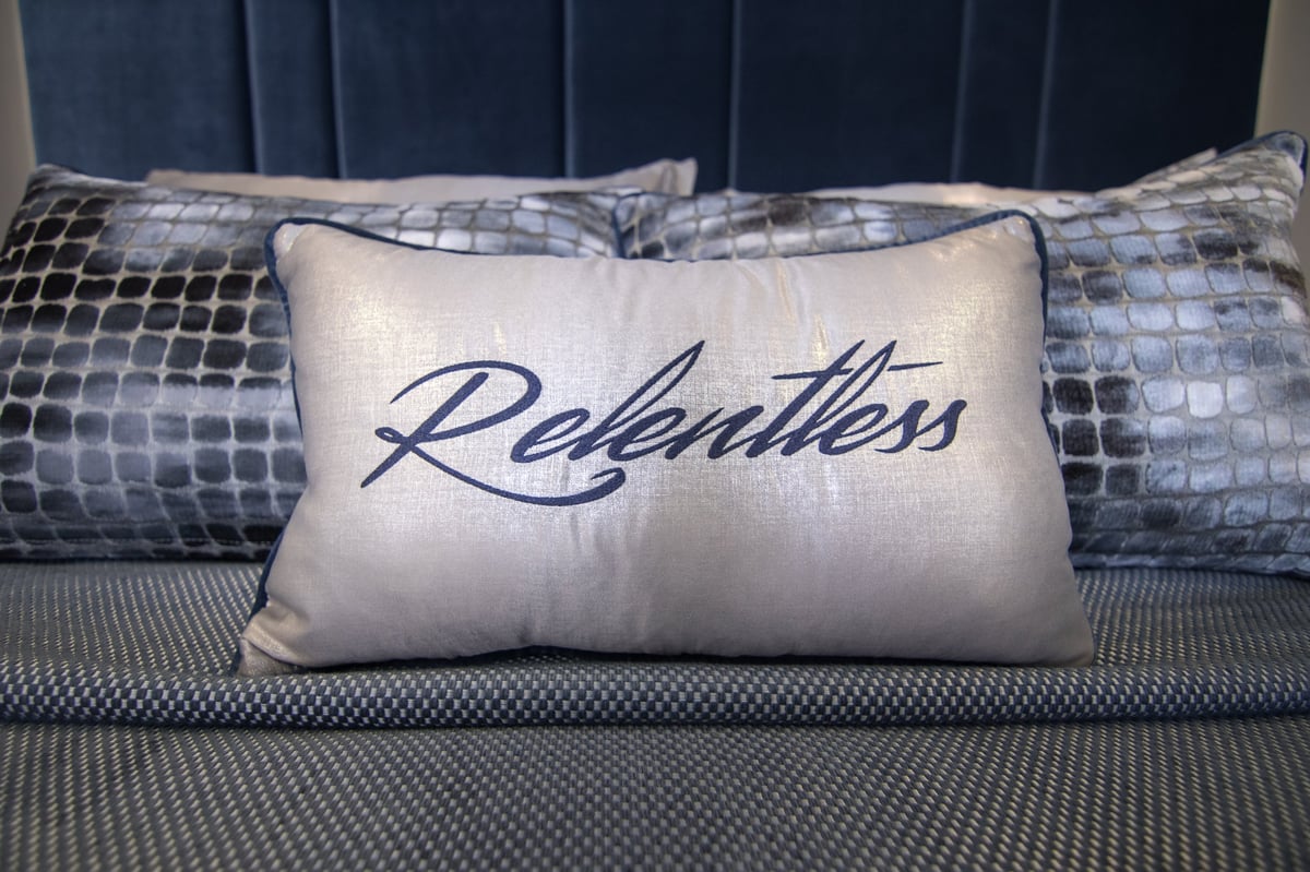 Cream-colored decorative pillow with Relentless script text displayed on a bed with blue patterned pillows
