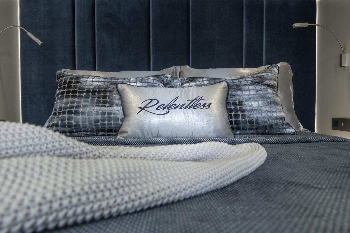 Modern bedroom with neatly made bed featuring gray textured pillows and a white decorative pillow with Relentless script, dark headboard, and folded gray blanket in foreground
