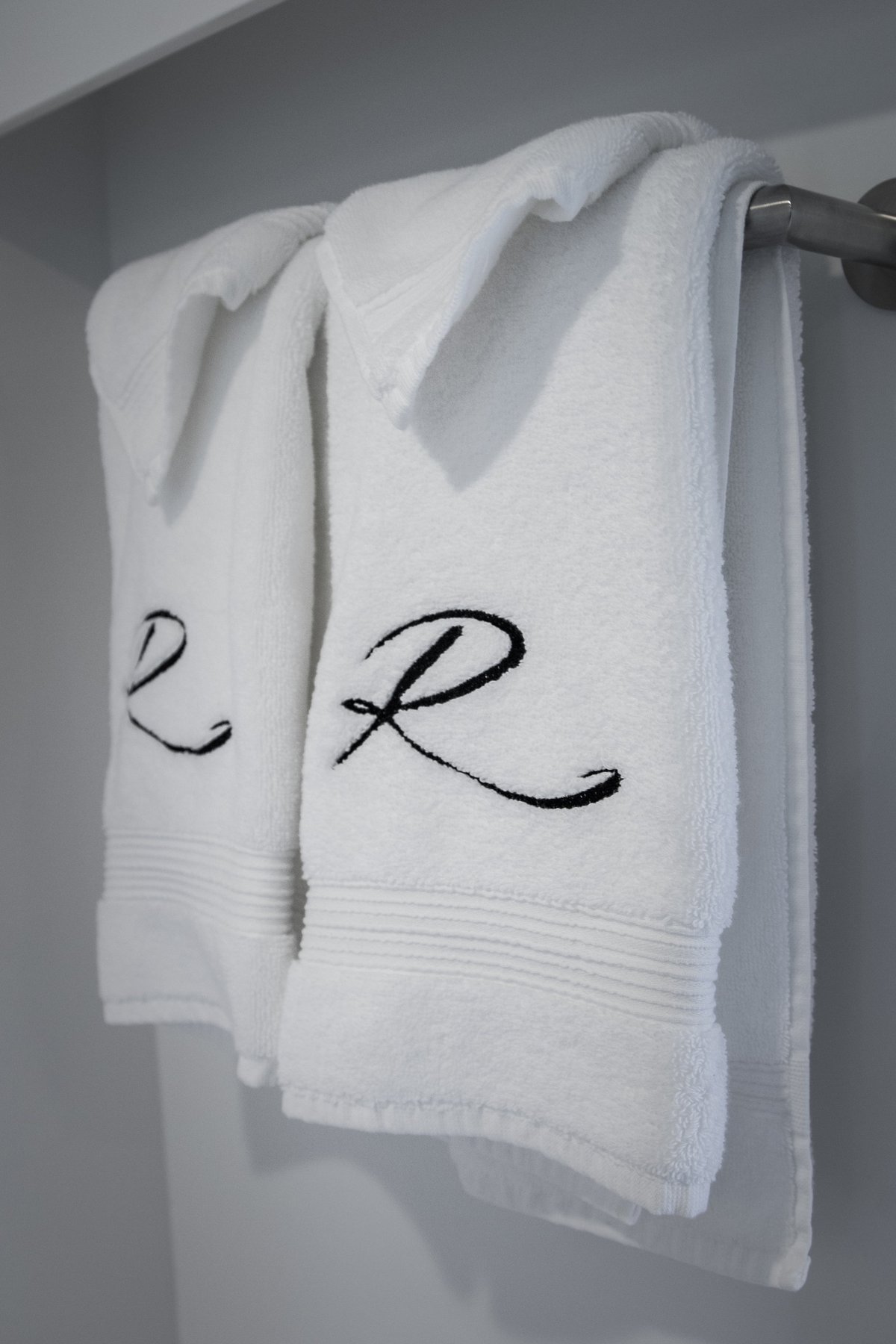 Two white embroidered towels with letters L and R hanging on a dark towel rod against a gray wall