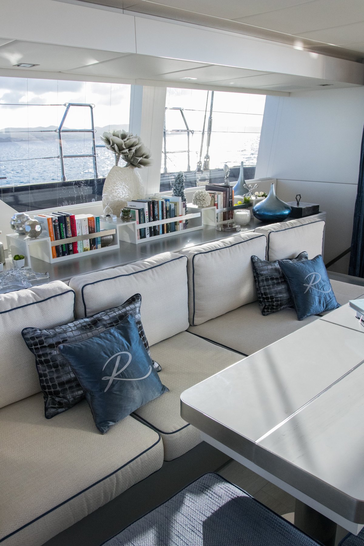 Modern yacht cabin with white seating, blue Relentless monogram throw pillows, shelving with books and decor, and ocean views through windows