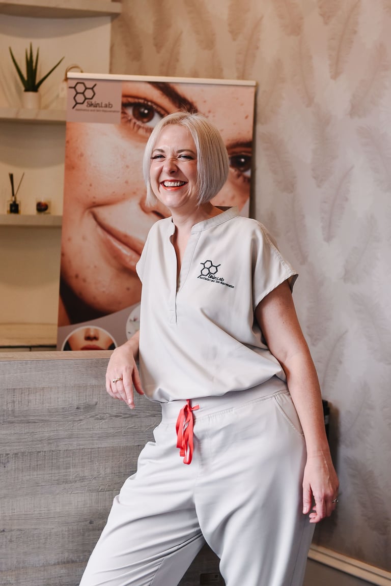 Professional woman with blonde hair in white spa uniform sitting in a modern salon interior with skincare branding on the wall behind her