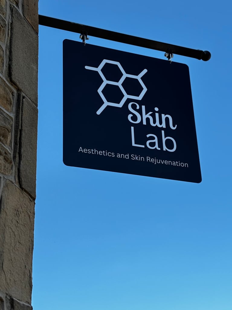 A dark blue hanging sign for Skin Lab with a molecular hexagon logo and subtitle Aesthetics and Skin Rejuvenation mounted on a brick building against a clear blue sky