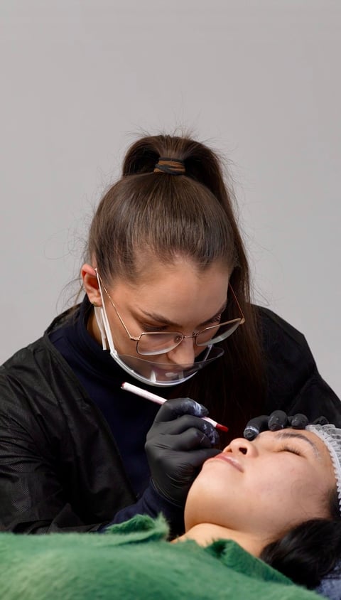 Permanent makeup artist wearing protective eyewear and gloves applying microblading to a clients eyebrow
