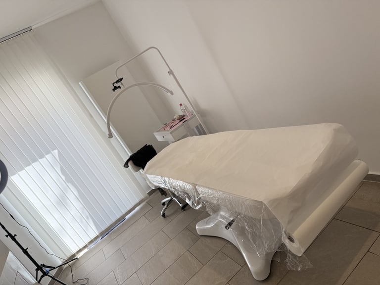 Minimalist treatment room with white padded chair covered in plastic, mirror, and tall window with blinds