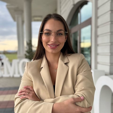 Professional woman wearing beige blazer and glasses smiling at camera with arms crossed in modern office setting with windows