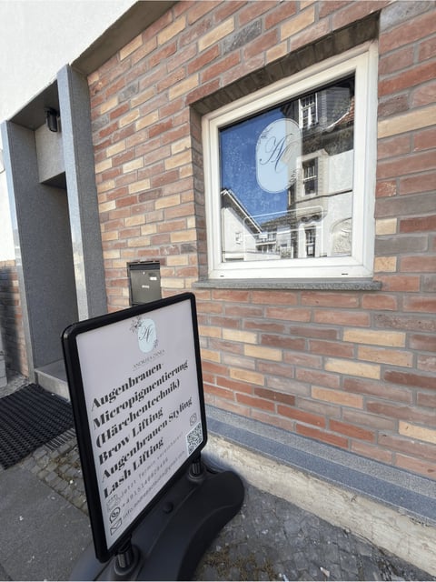 Brick storefront with white-framed window displaying blue signage and a rolling promotional sign on the ground