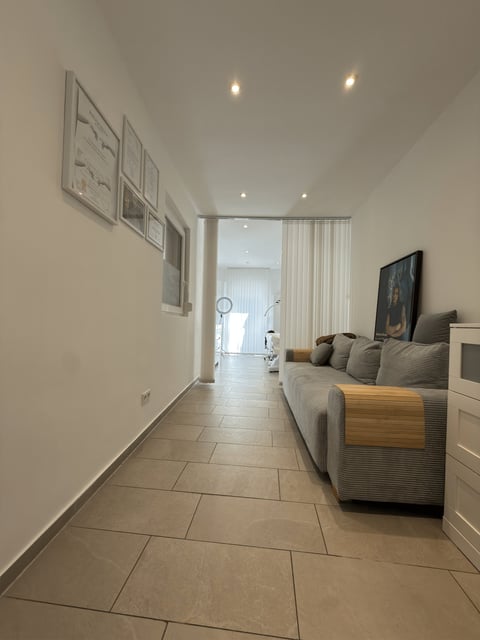 Modern living room hallway with gray upholstered sofa, wood accents, tiled flooring, and framed artwork on walls