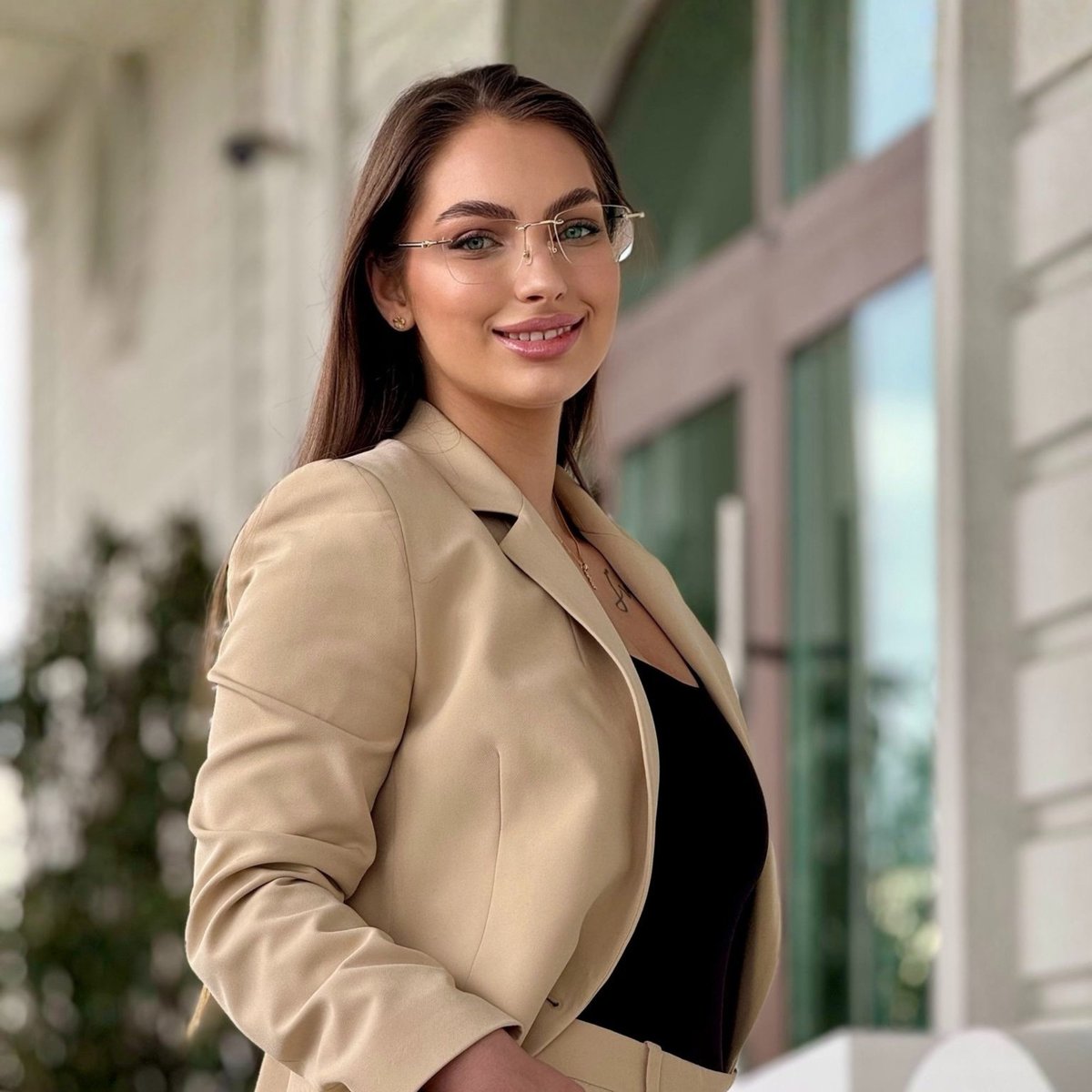 Professional woman wearing glasses and a beige blazer over black top, smiling at camera in front of modern building with large windows