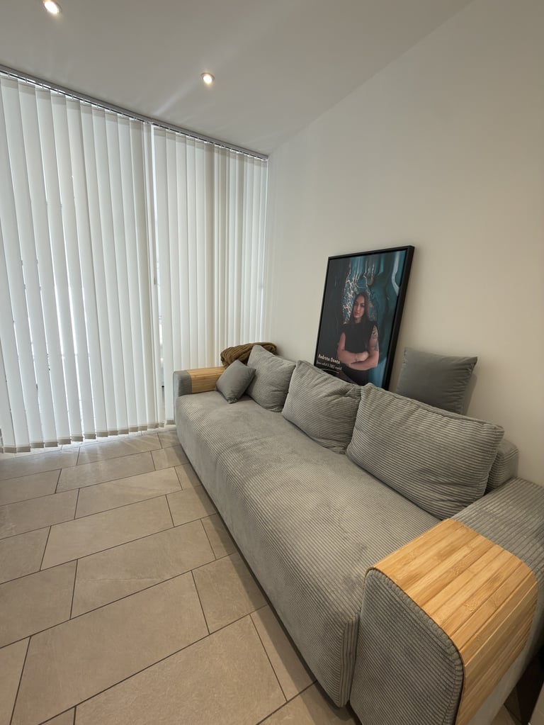 Modern living room with a gray sectional sofa, wooden side table, portrait artwork on wall, and vertical blinds beside a window