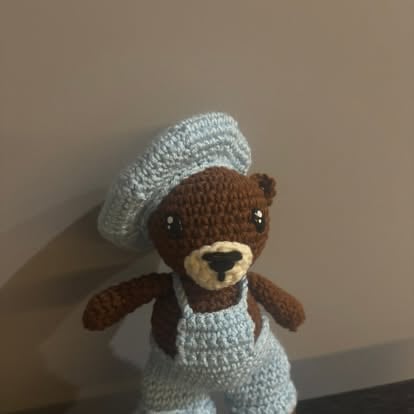 Brown crocheted teddy bear wearing a gray knit beret and bib against a beige background